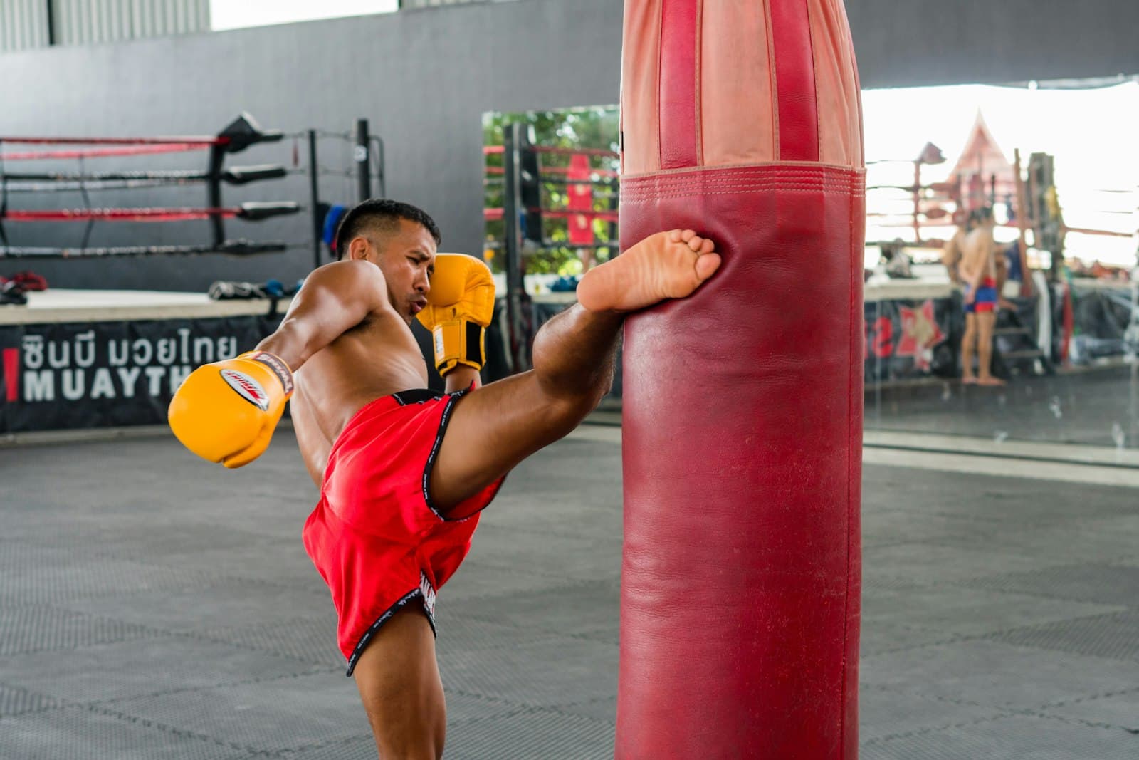 A Muay Thai fighter in red Thai shorts kicking a heavy bag