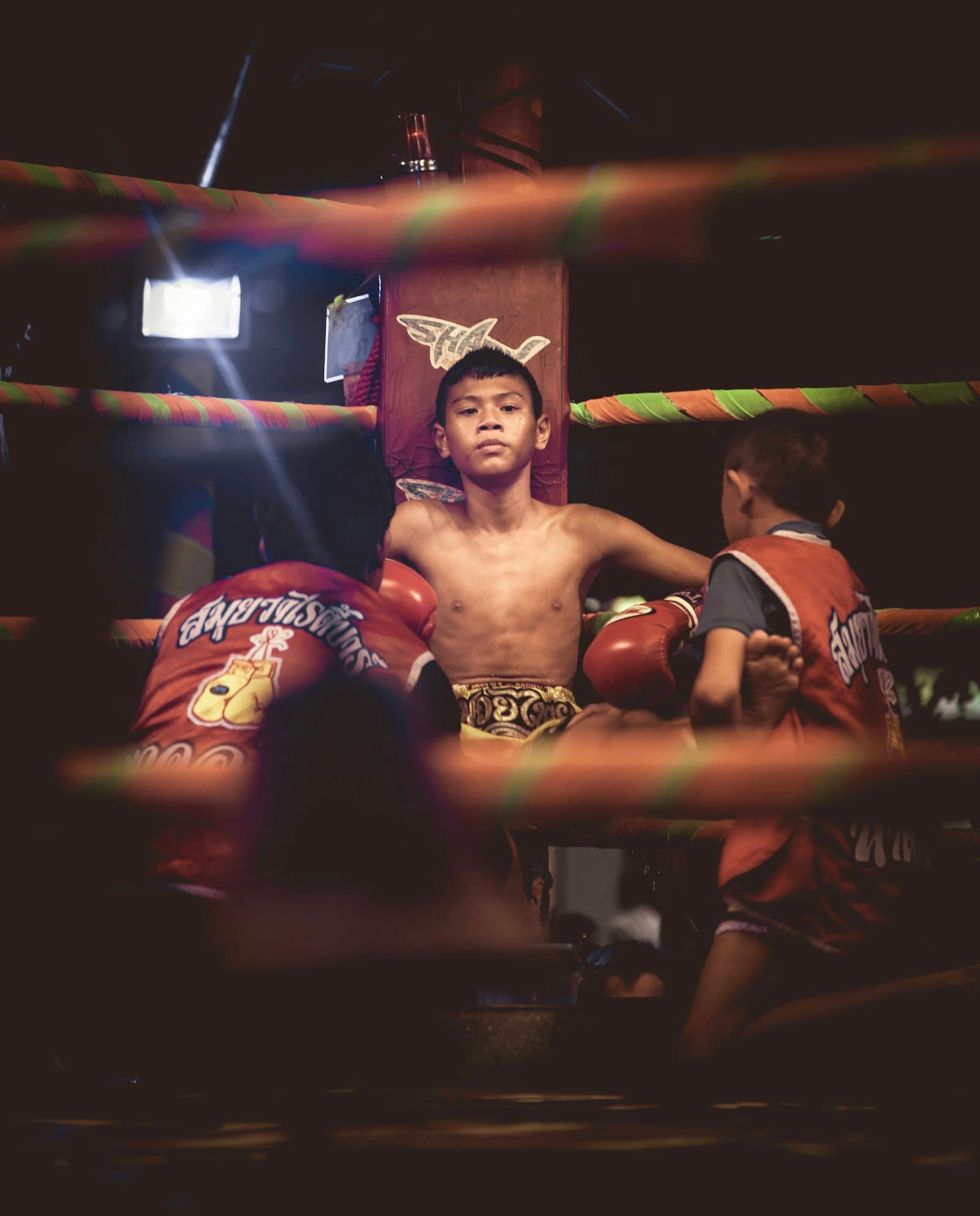 A young fighter wearing a mongkol headband in the ring corner, between trainers